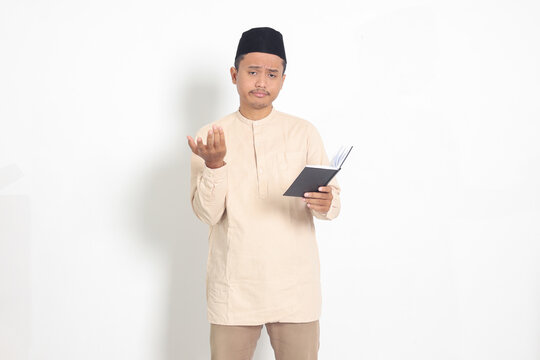 Portrait of confused Asian muslim man in koko shirt with peci difficulty understanding the contents of the book, reading a textbook. Isolated image on white background