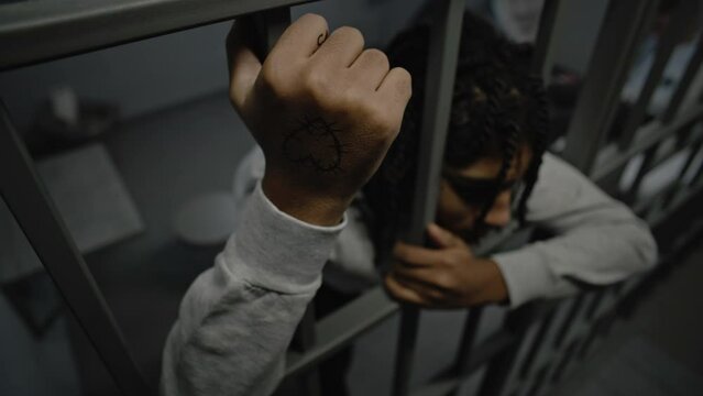 Depressed African American teenage criminal stands in prison cell in jail, holds metal bars. Young prisoners sit on bed in background. Juvenile detention center or correctional facility. High angle.