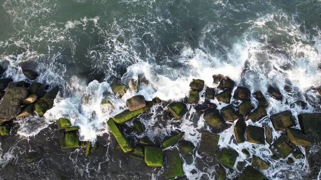 Aerial view of waves crashing against a rocky, moss-covered breakwater, depicting the powerful beauty of nature, ideal for environmental or marine-themed projects