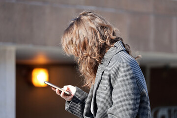 Young woman in coat with smartphone in hands. Lady using mobile phone on a city street in spring