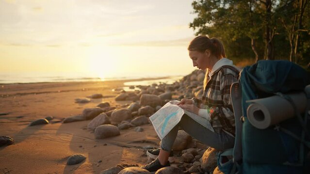 Tourist woman looking a way on sea beach. Female having break in hike finding route look at paper map admiring nature at sunset sitting on stones. Camping hiking tourism travel adventure concept.