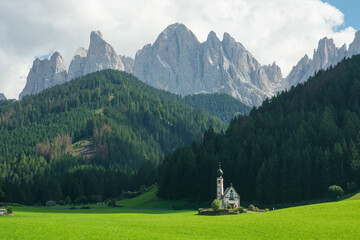 View of the Church of St. Johann is in a beautiful scenic location against the Geisler peaks covered with clouds in Santa Maddalena village, Val Di Funes, Italy - 764554173