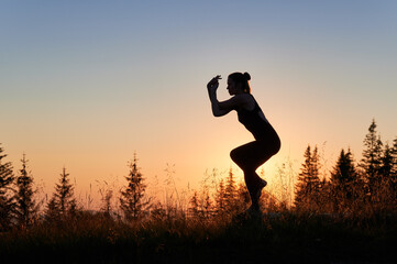 Elegant silhouette of girl doing eagle pose. Young woman exersicing in mountains at dawn. Practicing yoga on fresh air.