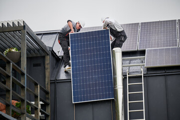 Men roofers installing solar panel system on roof of house. Technicians in helmets lifting up...