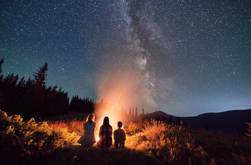 Young family stargazing together in mountains. Three girls warming up near bonfire. Family looking...