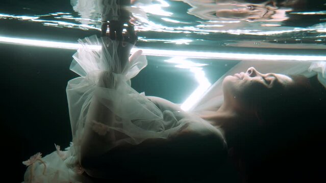 A lovely girl, in a tight-fitting white, swims gracefully under the water, like a fantastic mermaid, surrounded by neon reflections that add mystery to her appearance.