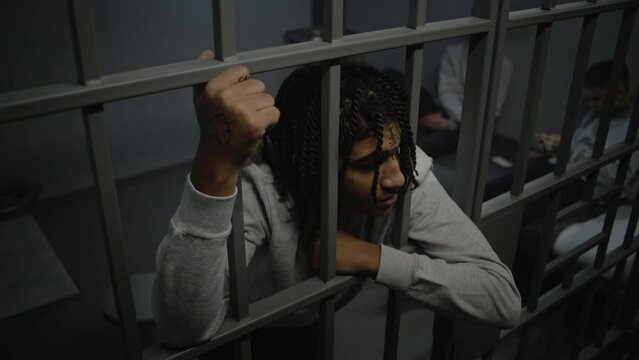 Angry African American teenage prisoner stands in prison cell in jail, holds metal bars. Young inmates play cards on bed in the background. Youth detention center or correctional facility. High angle.