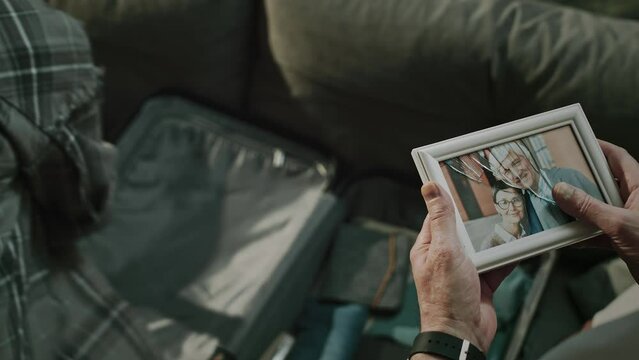 High angle shot of unhappy mature Caucasian couple separating after many years together. Wife packing suitcase and man looking at broken picture frame of them smiling at camera