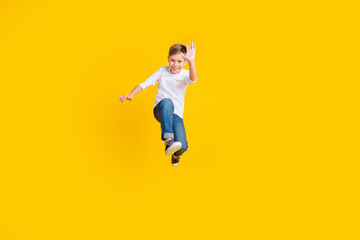Fototapeta na wymiar Full length photo of friendly cool boy wear white shirt jumping high waving arm hi empty space isolated yellow color background