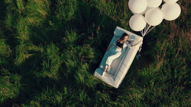 A girl with balloons licks on a couch standing in a field. drone footage of a woman in a field with a sofa and balloons. Concept of holiday, happiness, solitude.