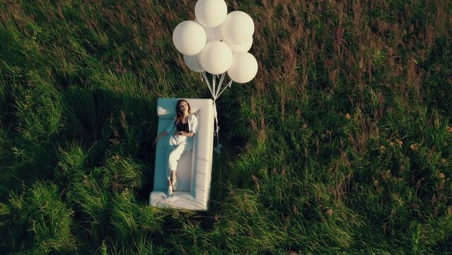 A girl with balloons licks on a couch standing in a field. drone footage of a woman in a field with a sofa and balloons. Concept of holiday, happiness, solitude.