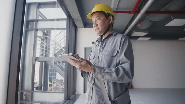 Tilt up portrait of adult Asian man in hardhat and gray uniform checking digital tablet at construction site and looking at camera