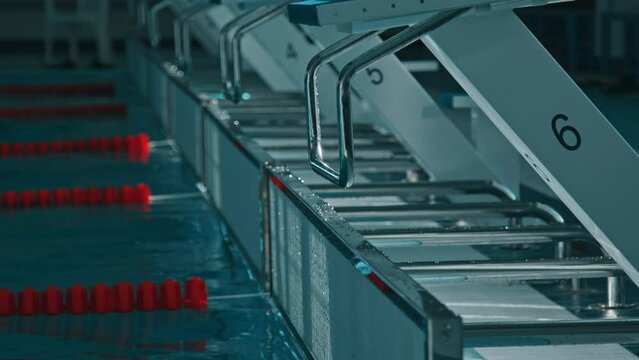 Close-up no people shot of row of dive blocks at deep end of empty swimming pool, red lane dividers bobbing on surface, and offscreen swimmer splashing in blue water