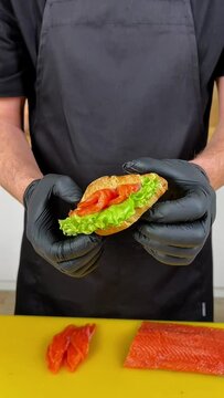 Chef in black apron and gloves presents a salmon sandwich above a yellow cutting board, depicting food preparation or culinary artsideal for cooking or healthy eating concepts