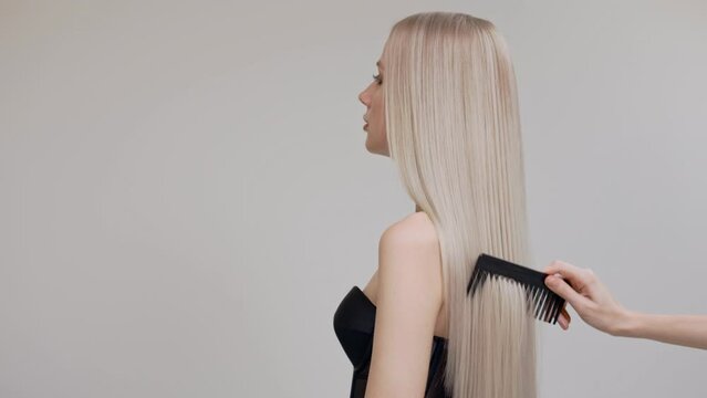 A blonde girl is combing her chic long straight hair in the studio on a light background.