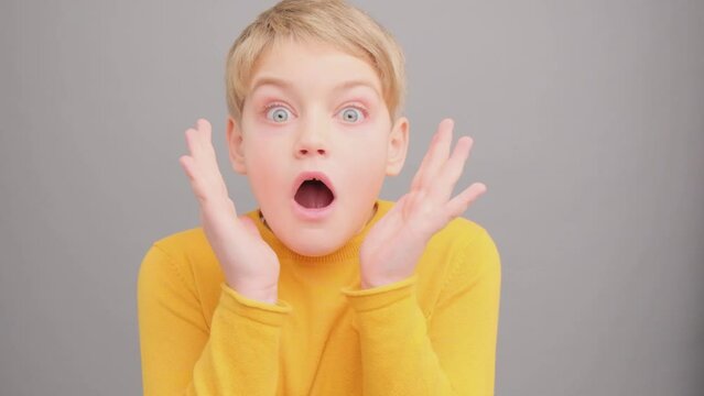 A little boy in a yellow T-shirt looks at the camera and is surprised, in a studio on a gray background. Child 8 years old.
