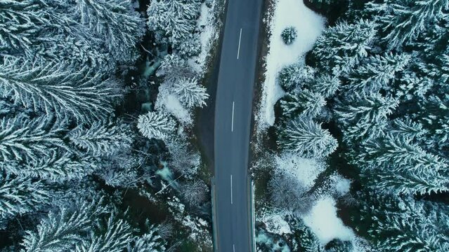Aerial shot of drone fly over empty asphalt road in snow covered forest in the mountains. Cinematic road trip in cold winter conditions. 