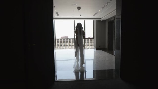 A bride in a white pantsuit and veil walks down a dark corridor to the window.