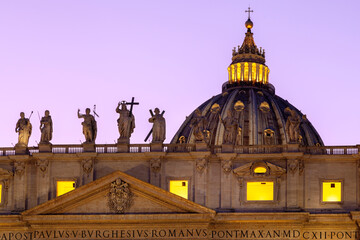 Details st at the rooftop of San Pietro Basilica (Saint Peter Cathedral) of Vatican City during dusk time, with statues and the illuminated dome of the cathedral.