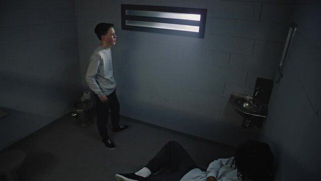 Diverse teenagers talk while serving term in jail. Young Caucasian prisoner stands near the window in prison cell, African American inmate sits on bed. Juvenile detention center. Surveillance camera.