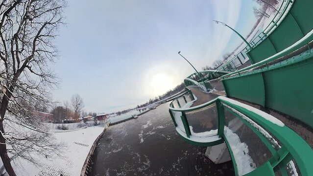 This is a 360 -degree video of a pedestrian bridge in winter. The bridge is made of metal and painted green. It has a curved design and is surrounded by snow-covered trees and bushes. The bridge is