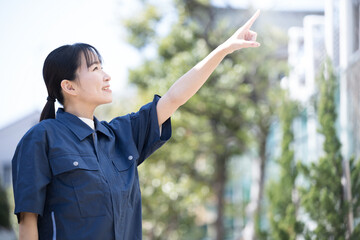 新緑の青空の屋外で作業着を着た女性　未来や目標を目指して指を差す