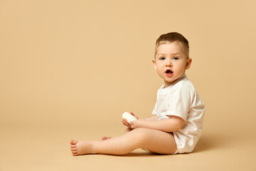 Cute little baby, boy, child in diaper and t-shirt calmly sitting on bed with body cream isolated over beige studio background. Concept of childhood and family, care, parenthood, infancy and heath