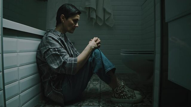 Lowkey shot of adult Caucasian woman sitting on floor in cold dark bathroom with her knees to chest while hiding from abusive husband during domestic violence episode