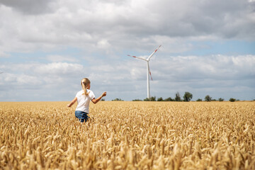 Back view little cute happy blond caucasian girl enjoy walking ripe wheat ear field against wind mill turbine farm on warm sunny summer day. Child freedom future clean energy environment concept - Powered by Adobe