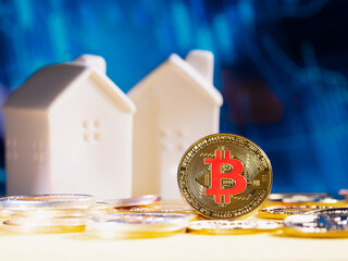 Bitcoin coin stands against a blurred blue background with financial graphs and two white model houses in the background. cryptocurrency and real estate. finance and investment on stock market
