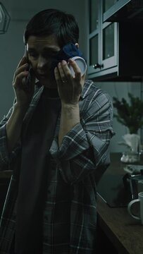 Vertical medium shot of battered Caucasian woman holding ice compress on her bruised face and making phone call for help, living with domestic tyrant in cold dark apartment