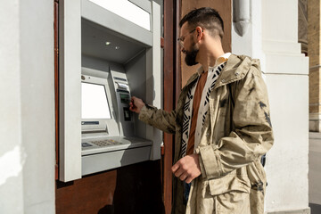 Young fashionable hipster backpacker traveler man using credit card to withdraw money from ATM...