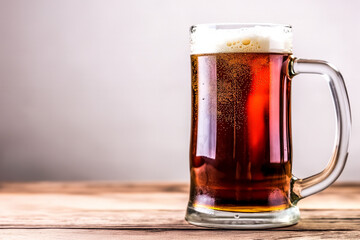 A glass mug of dark beer sits on a wooden table. The beer has a thick head of foam floating on top. The glass has a handle and a spout. - obrazy, fototapety, plakaty