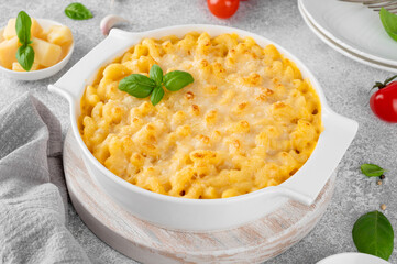 Mac and cheese in a white baking dish on a wooden board on a gray concrete background. American cuisine, comfort food. Copy space.