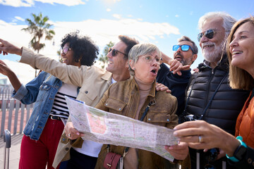 Portrait of mature diverse tourist friends holding a map on city street enjoying happy holidays. Mid aged people looking and pointing places of interest tourism. Traveling discussing direction to take