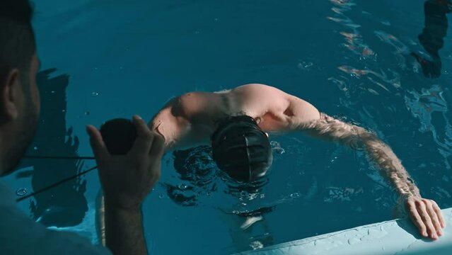Medium over-shoulder shot of competitive young male athlete in black cap and swimming goggles looking at stopwatch, shaking head, dipping face in water and talking to coach with desperation and dismay