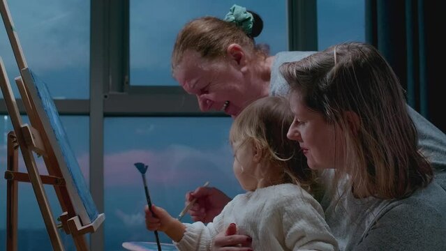 An elderly art teacher explains a technique to a toddler, painting on canvas with oil colors. Three-generation concept, Grandmother teaches granddaughter how to paint a draw