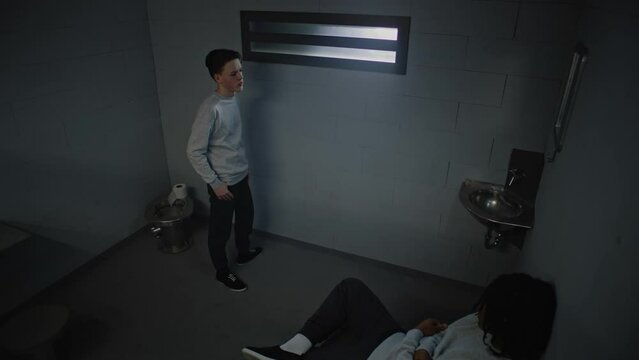 Two multiethnic teenagers talk serving imprisonment term for crimes in jail. One young criminal walks around the prison cell, another inmate sits on the bed. Youth detention center. CCTV camera angle.