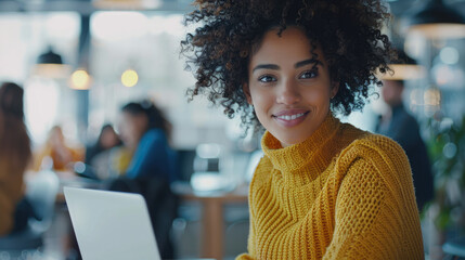 A cinematic shot of an attractive woman with curly hair wearing a yellow sweater sitting at her desk in front of a laptop, smiling and looking into the camera  - obrazy, fototapety, plakaty