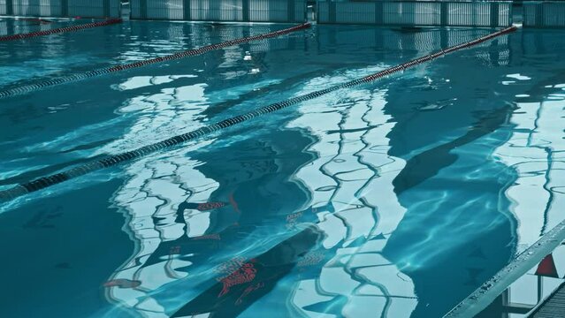 Full tilting down no people shot of dive blocks and rippling surface of turquoise water at deep end of swimming pool, with distorted reflections of windows and red letters on scoreboard, at daytime