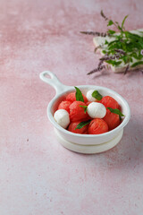 Fruit salad with watermelon balls and mozzarella cheese in white ceramic bowl on light rose background