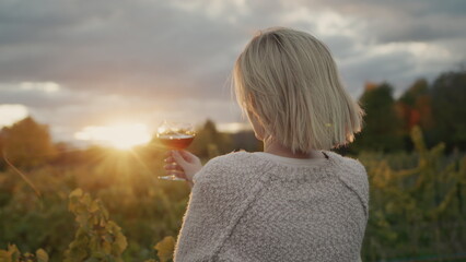 A woman with a glass of red wine in her hand stands against the background of a vineyard where the sun sets beautifully
