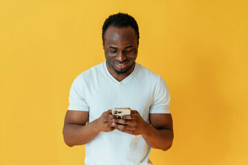 Holding smartphone and smiling. Black man is in the studio against yellow background