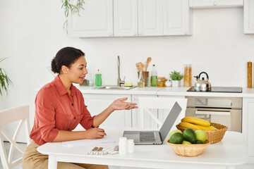 happy african american dietitian taking notes and giving online consultation on laptop from kitchen