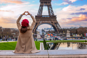 A woman with a french beret hat looks at the Eiffel Tower of Paris France, at forms a heart with her hands during sunset time