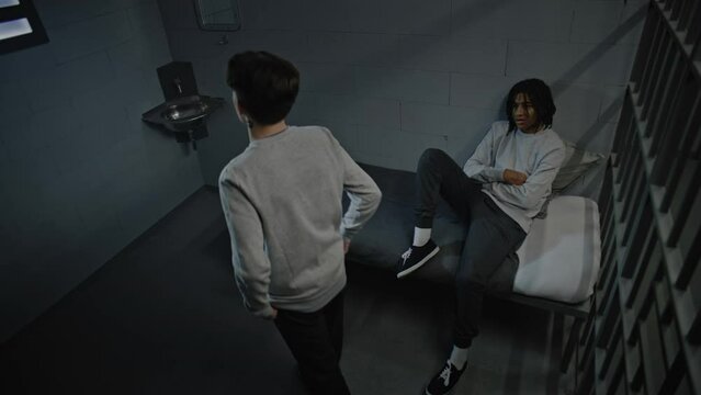 Two multicultural teenage criminals serve imprisonment term in jail. One walks around the prison cell, another lies on the bed. Youth detention center or correctional facility. Security camera angle.