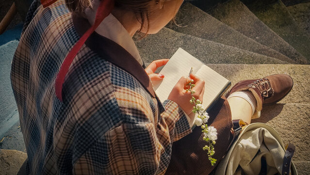A girl sits sideways on the steps of the university and writes in a notepad. She holds a blooming white branch in her hands. Retro image in the sun's rays