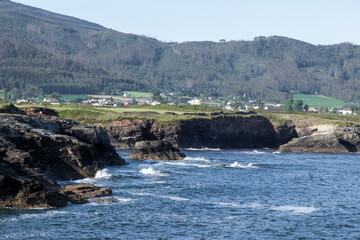 A coastal landscape with rocky shores, turbulent waters, and a green, hilly backdrop featuring a...