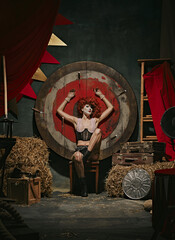 Woman in vintage costume and stage makeup poses before target over dark retro circus backstage...