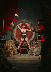 Young redhead woman with stage makeup and costume near target over dark retro circus backstage background. Man throwing knives. Concept of circus, theater, performance, show, retro and vintage